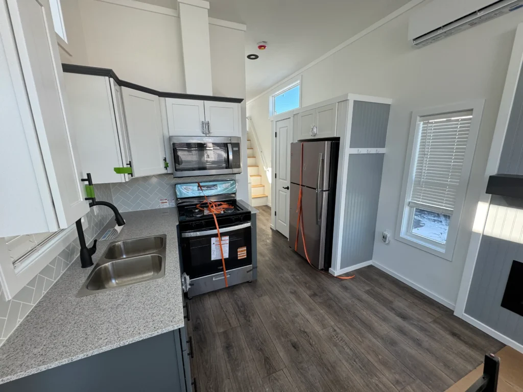 A complete tiny house kitchen configuration with a stainless steel stove, microwave, refrigerator, and granite counters.