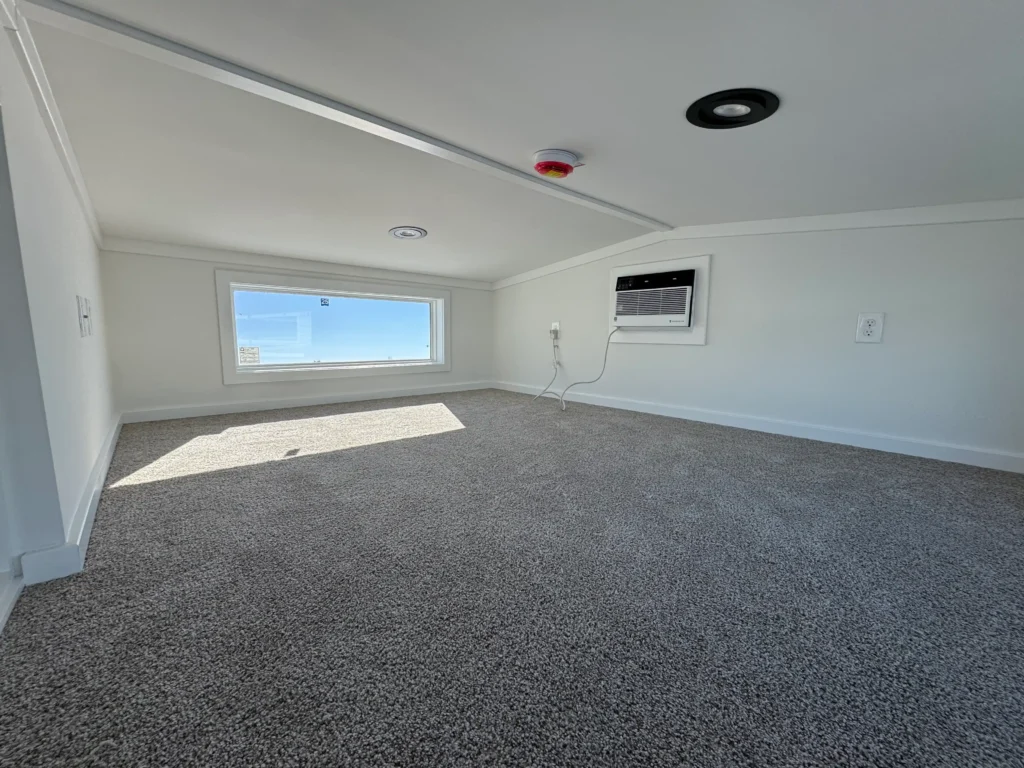 Carpeted upper loft sleeping area inside a park model cabin with a horizontal viewing window and a mini split AC.